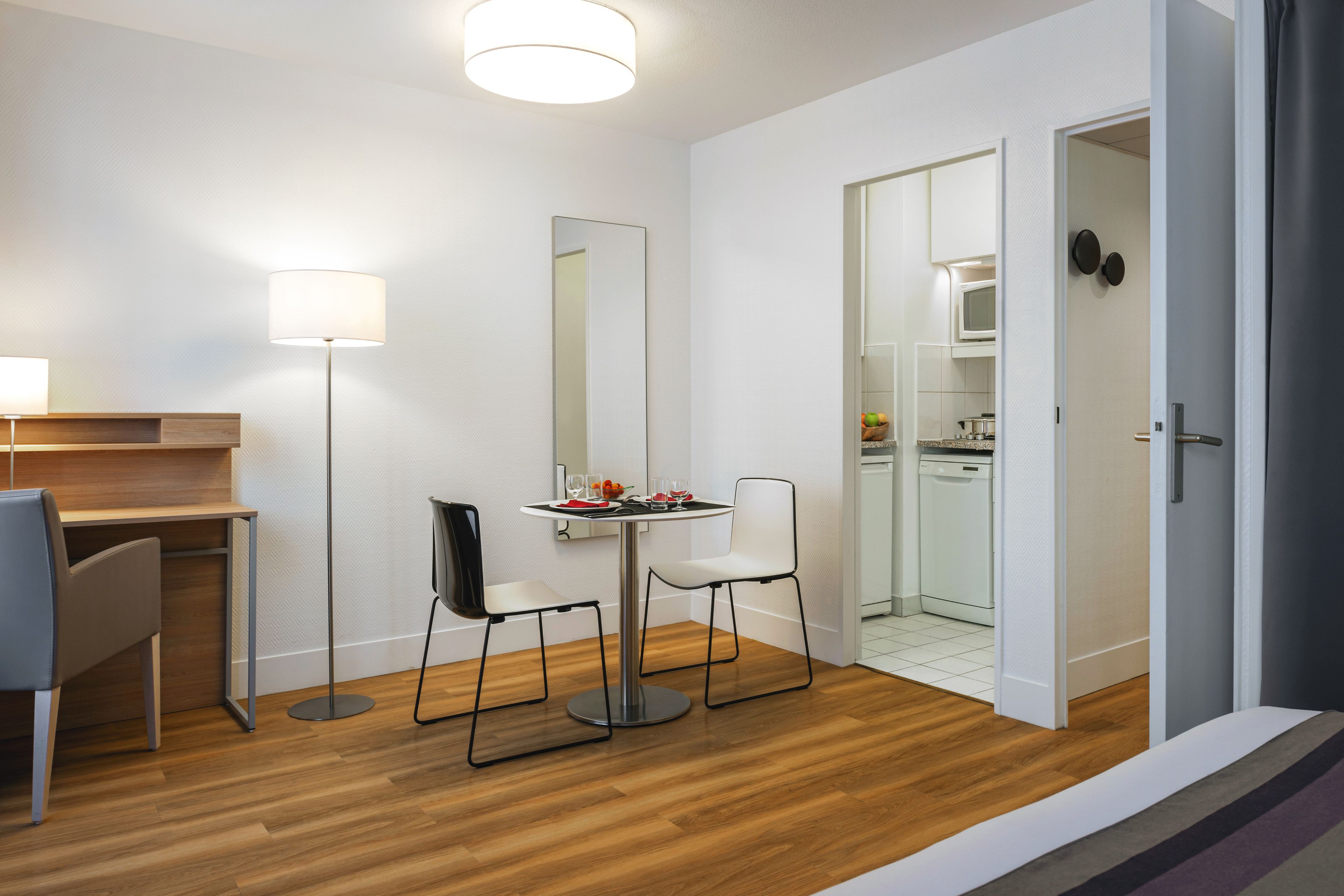 Habitación moderna y bien iluminada, con escritorio de madera, dos sillas, una mesa de comedor con lámpara y una cocina visible a través de una puerta abierta.