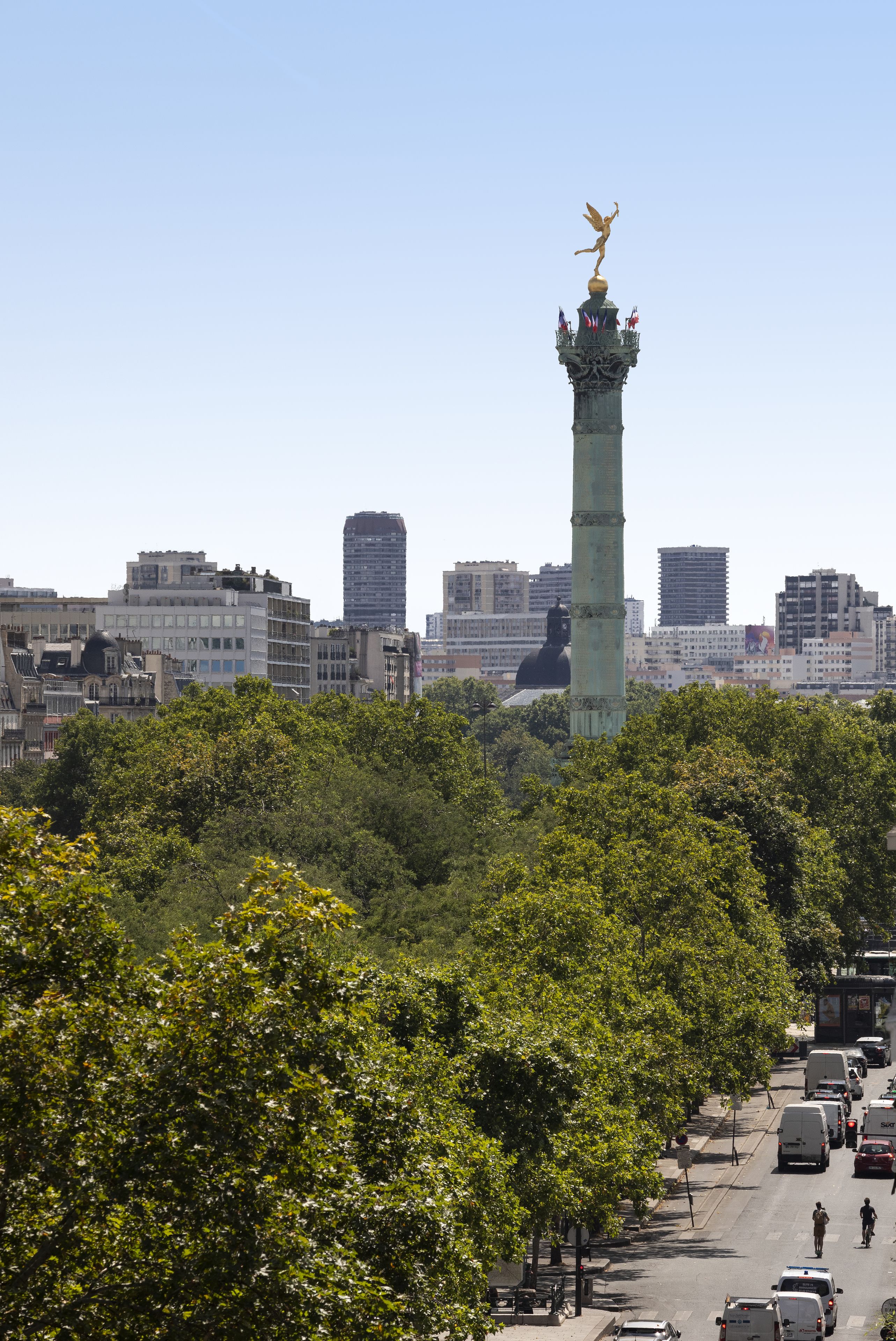 A cityscape with a prominent tower featuring a statue on top, surrounded by dense greenery and modern buildings.