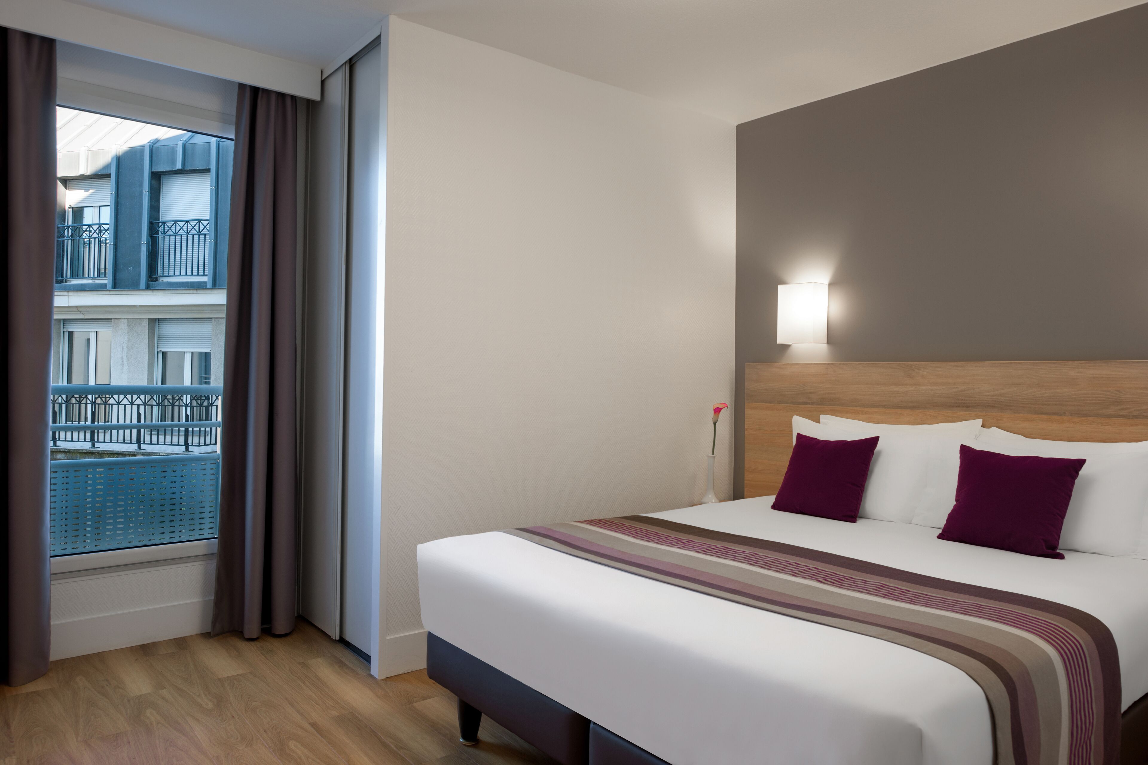 A hotel room with a large bed, a nightstand, a window with curtains, and a view of a building outside.
