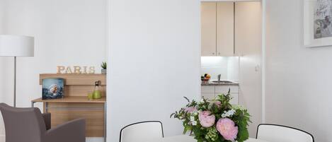 Ein moderner, minimalistischer Essbereich mit einem weißen Tisch, grauen Stühlen und einem Blumenstrauß. Im Hintergrund ist ein Holzregal mit einem ‚PARIS‘-Schild und einem gerahmten Bild zu sehen.