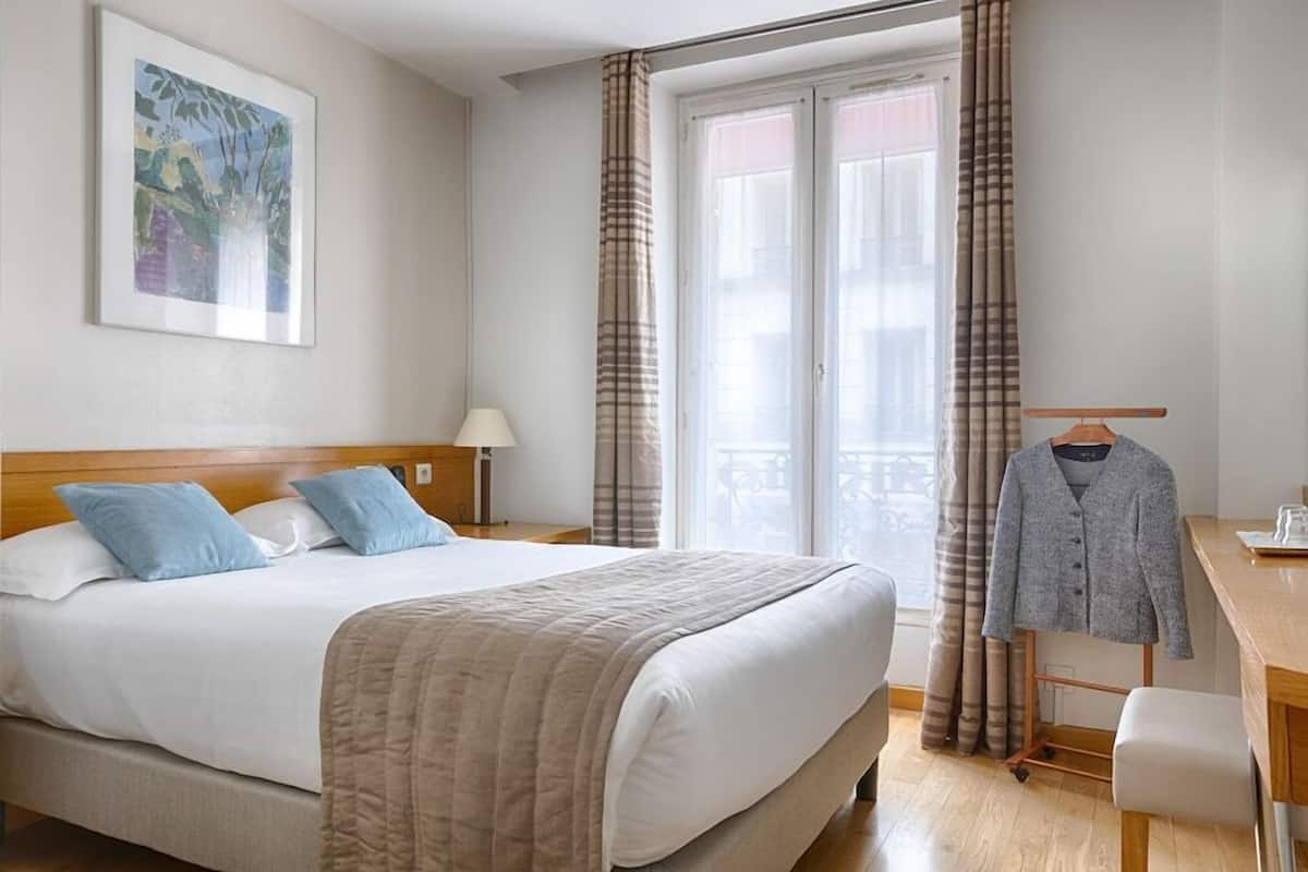 Ett hotellrum med en säng, ett skrivbord, en stol, en lampa och ett fönster med gardiner.