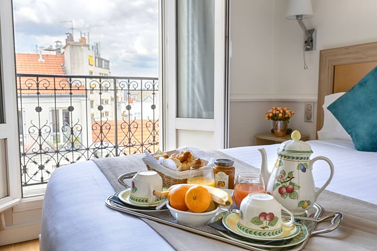 Ett välordnat frukostbricka med croissanter, apelsinjuice och en tekanna står på sängen i ett hotellrum med utsikt över byggnader genom ett fönster.