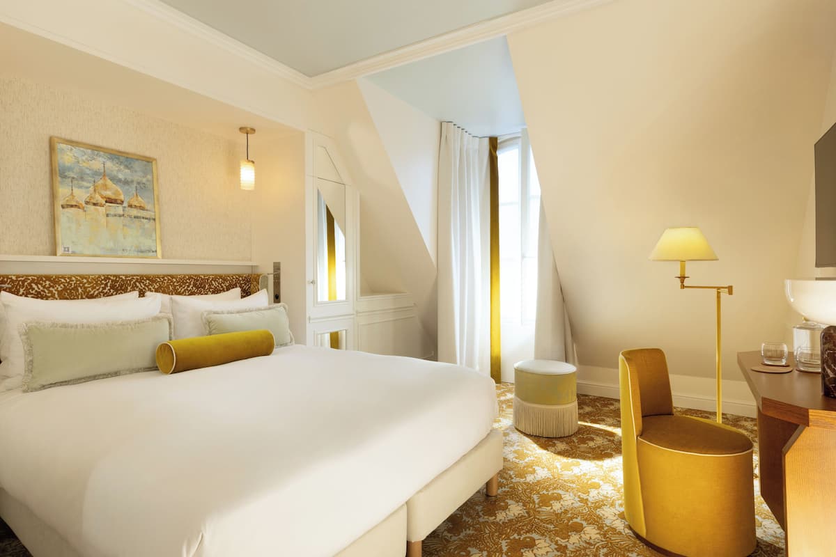 A hotel room with a large bed, a small round table, a chair, a lamp, and a painting on the wall.