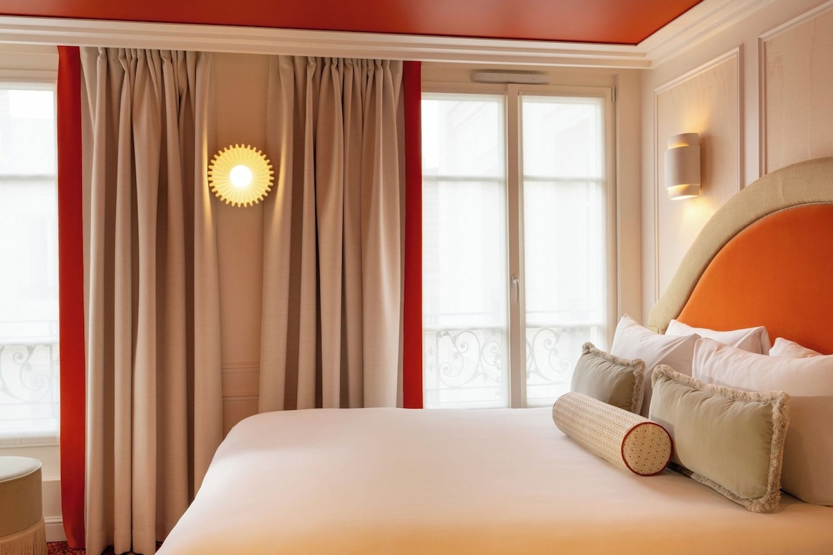 Une chambre d’hôtel avec un grand lit, des rideaux, une lampe fixée au mur et une fenêtre donnant sur un beau paysage.