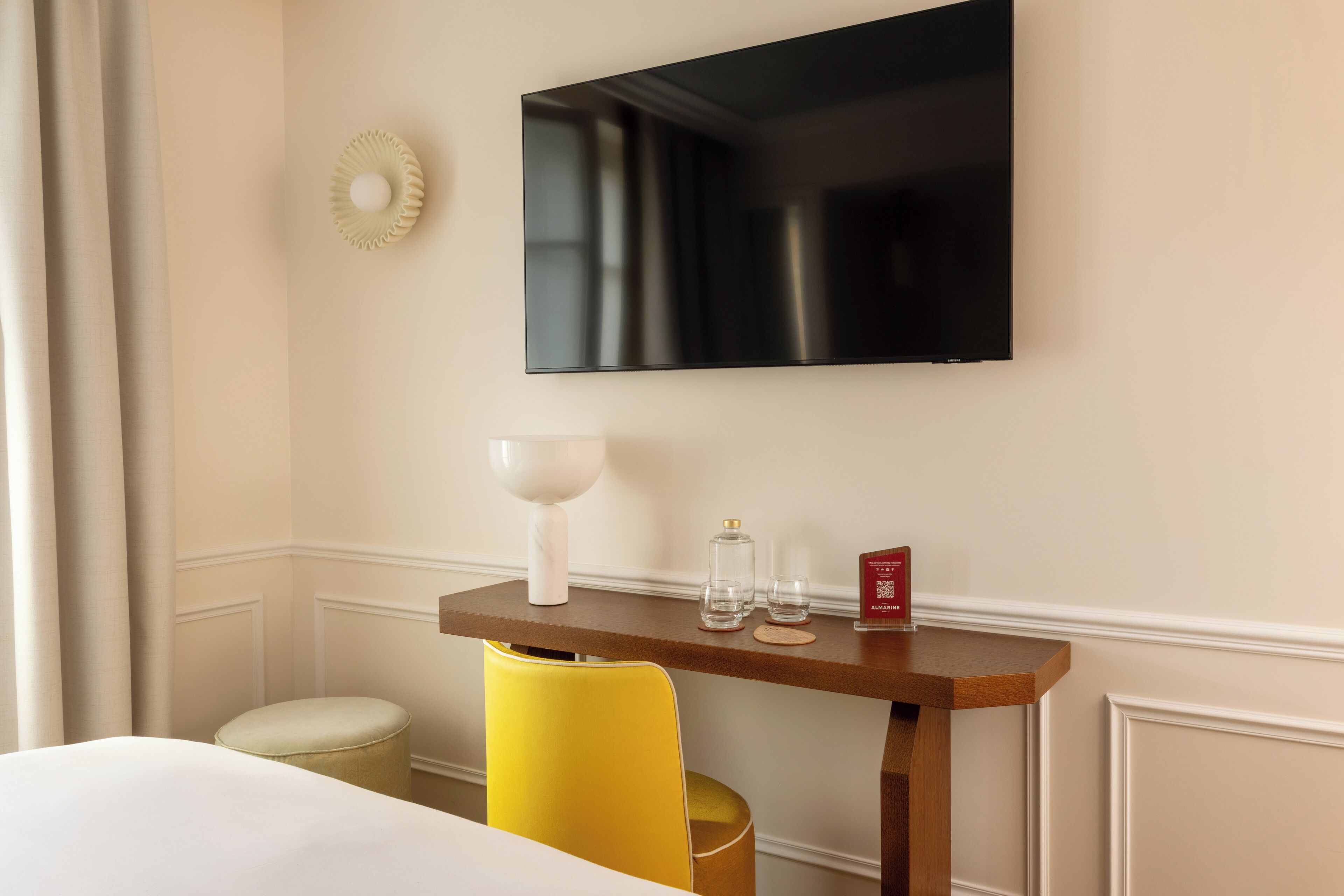 Et hotelværelse med en seng, en gul stol, et lille bord med en hvid lampe og et fladskærms-tv monteret på væggen.