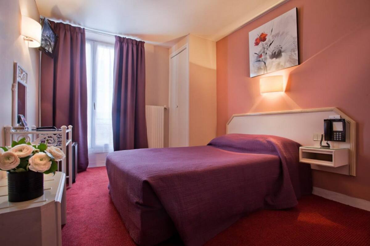 A hotel room with a bed, a desk with a lamp, a telephone, and a painting on the wall.