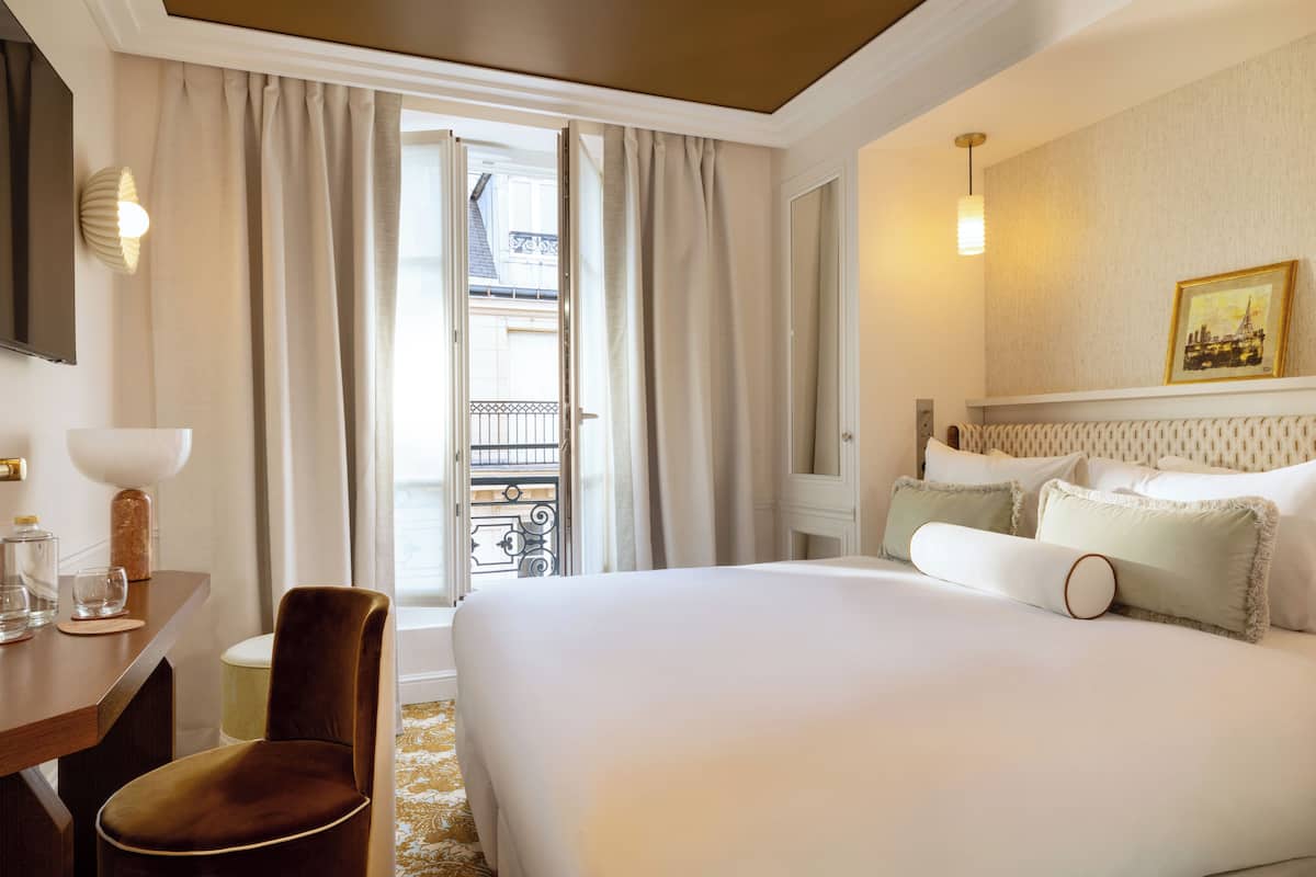 A hotel room with a large bed, a desk with a chair, a small table with glasses and a candle, and a balcony with a railing.