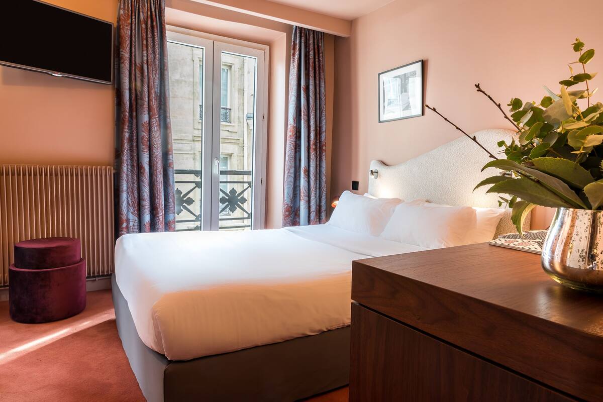 Habitación de hotel con cama, televisión, ventana con cortinas, radiador y una planta sobre una mesita de noche.