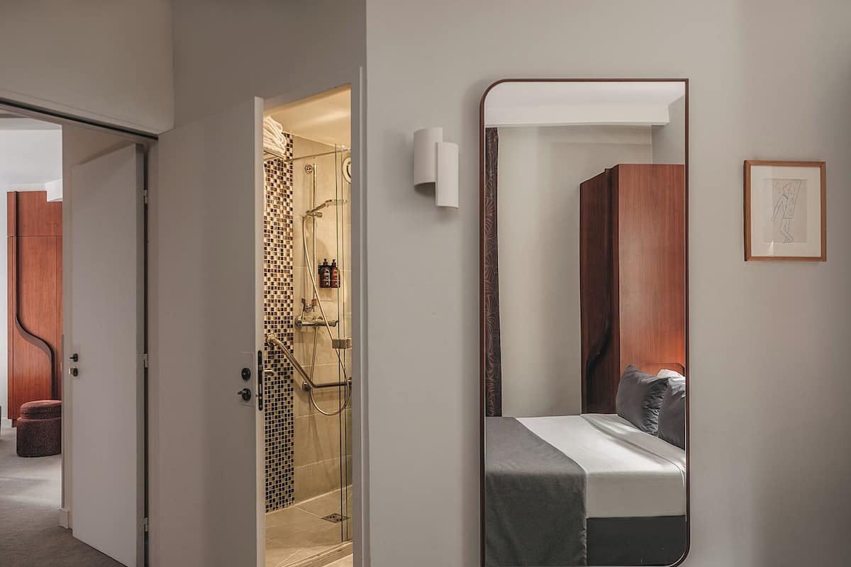 Una habitación de hotel moderna con un espejo grande que refleja una cama bien hecha, una zona de ducha con puerta de cristal y un armario de madera.