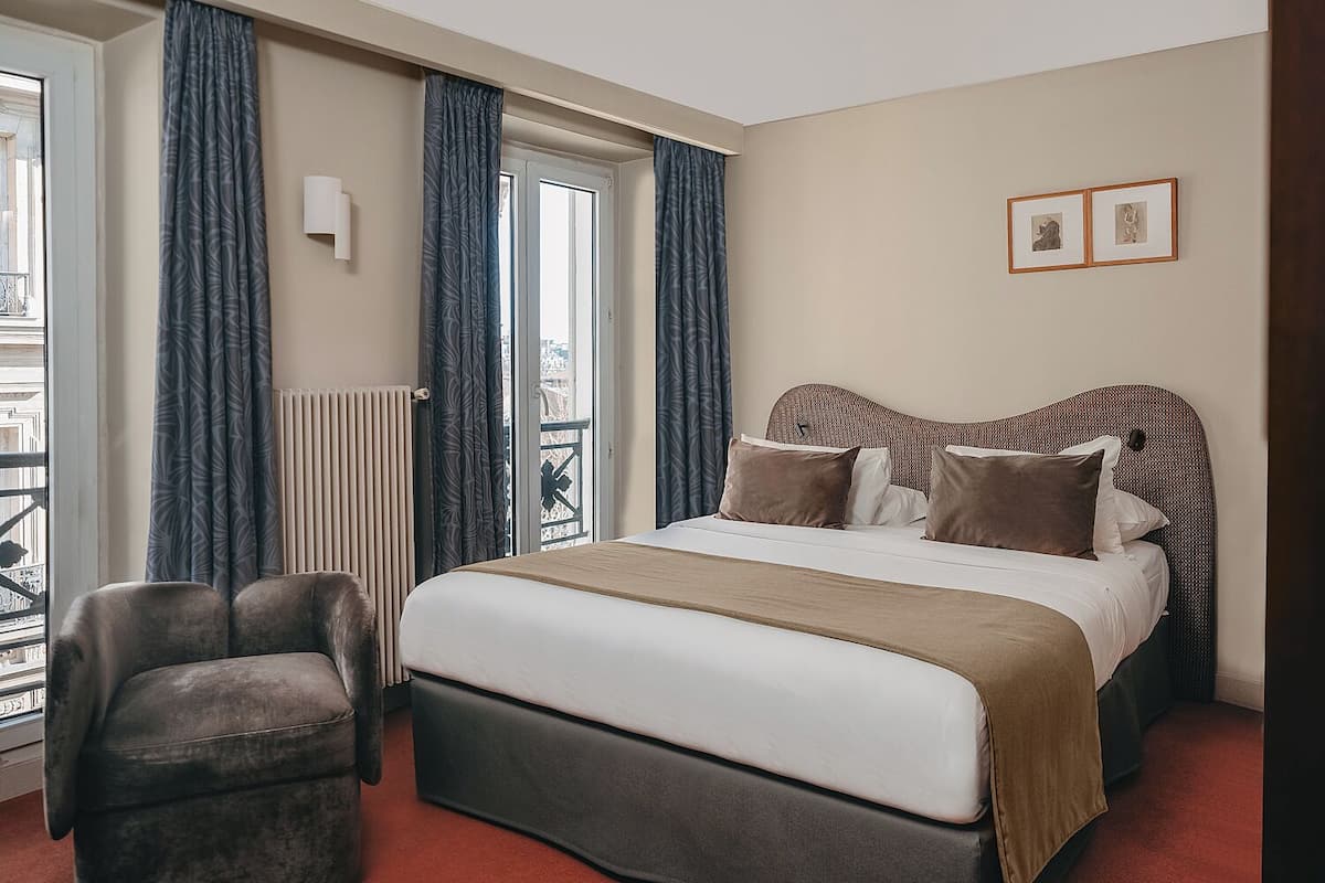 Habitación de hotel con una cama grande, una silla, un radiador y vistas a la ciudad a través de la ventana.