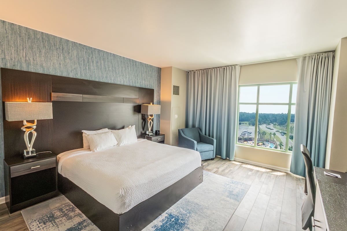 A modern hotel room with a large bed, two bedside tables with lamps, a chair, and a large window with curtains.