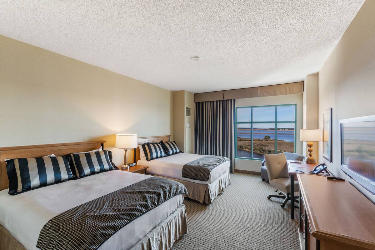 A hotel room with two beds, a desk, a chair, a TV, and a large window with a view.