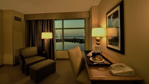 A hotel room with a view of a lake, a desk with a telephone, a chair, an ottoman, and a tray with food and a drink. - Hollywood Casino Gulf Coast (Bay St Louis)