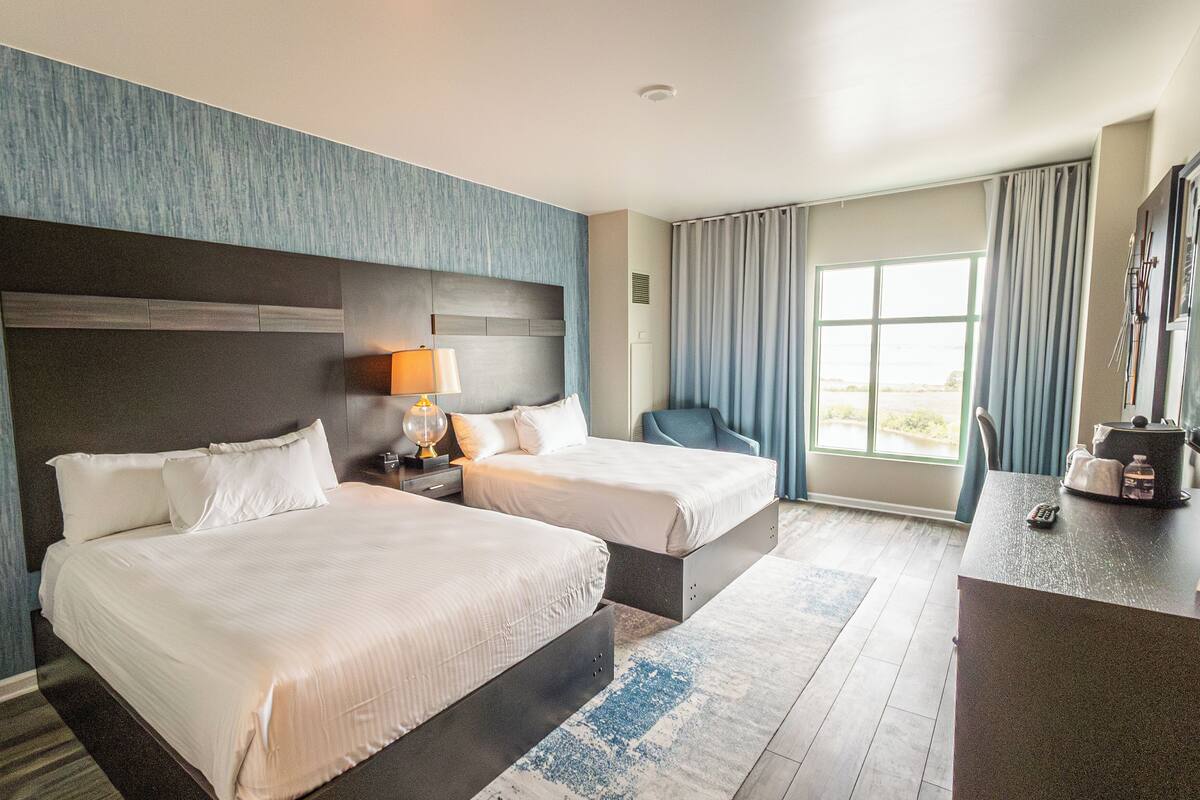 A hotel room with two beds, a desk, a chair, and a large window with curtains.