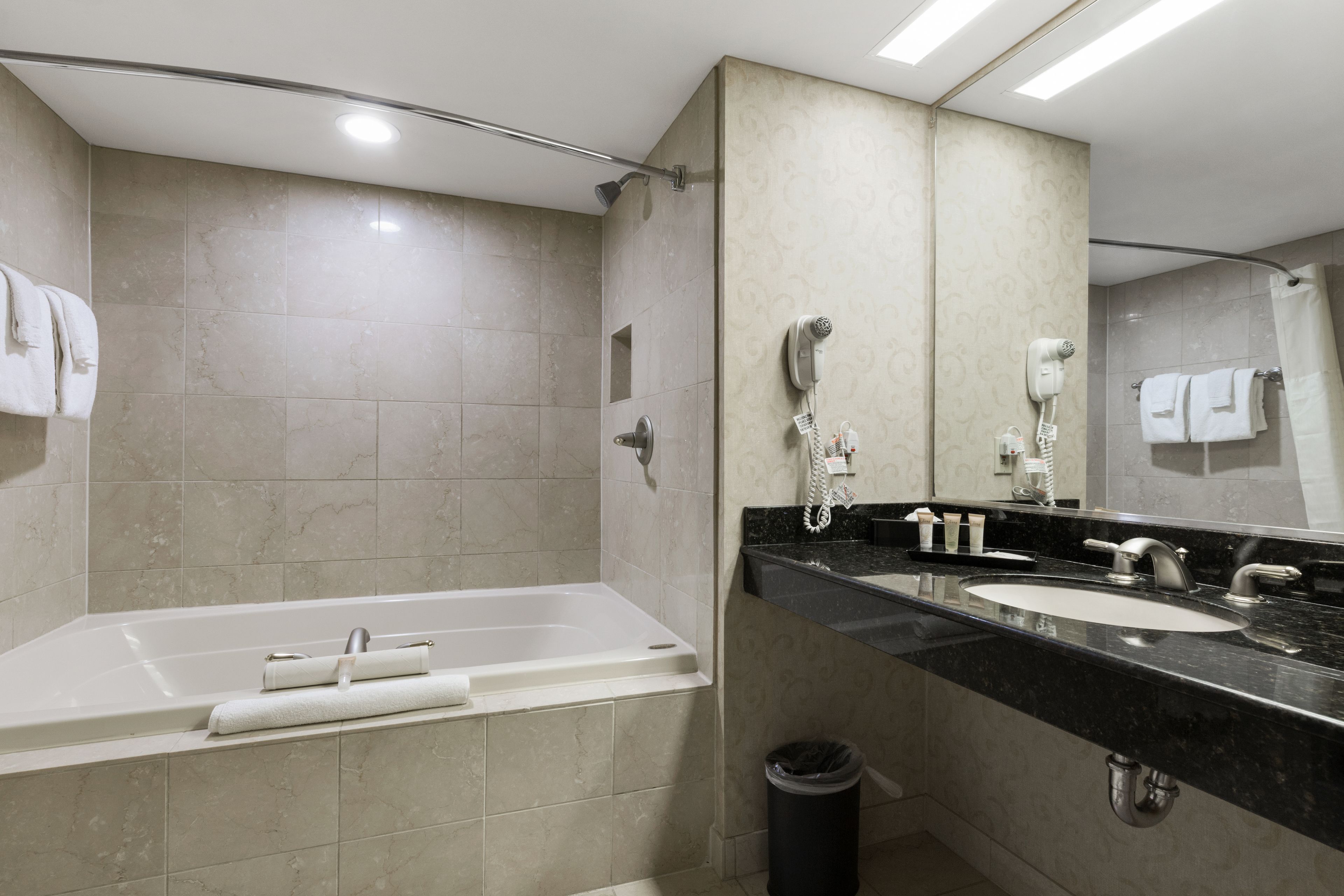 A modern bathroom with a bathtub, two sinks, a hairdryer, and a trash can.