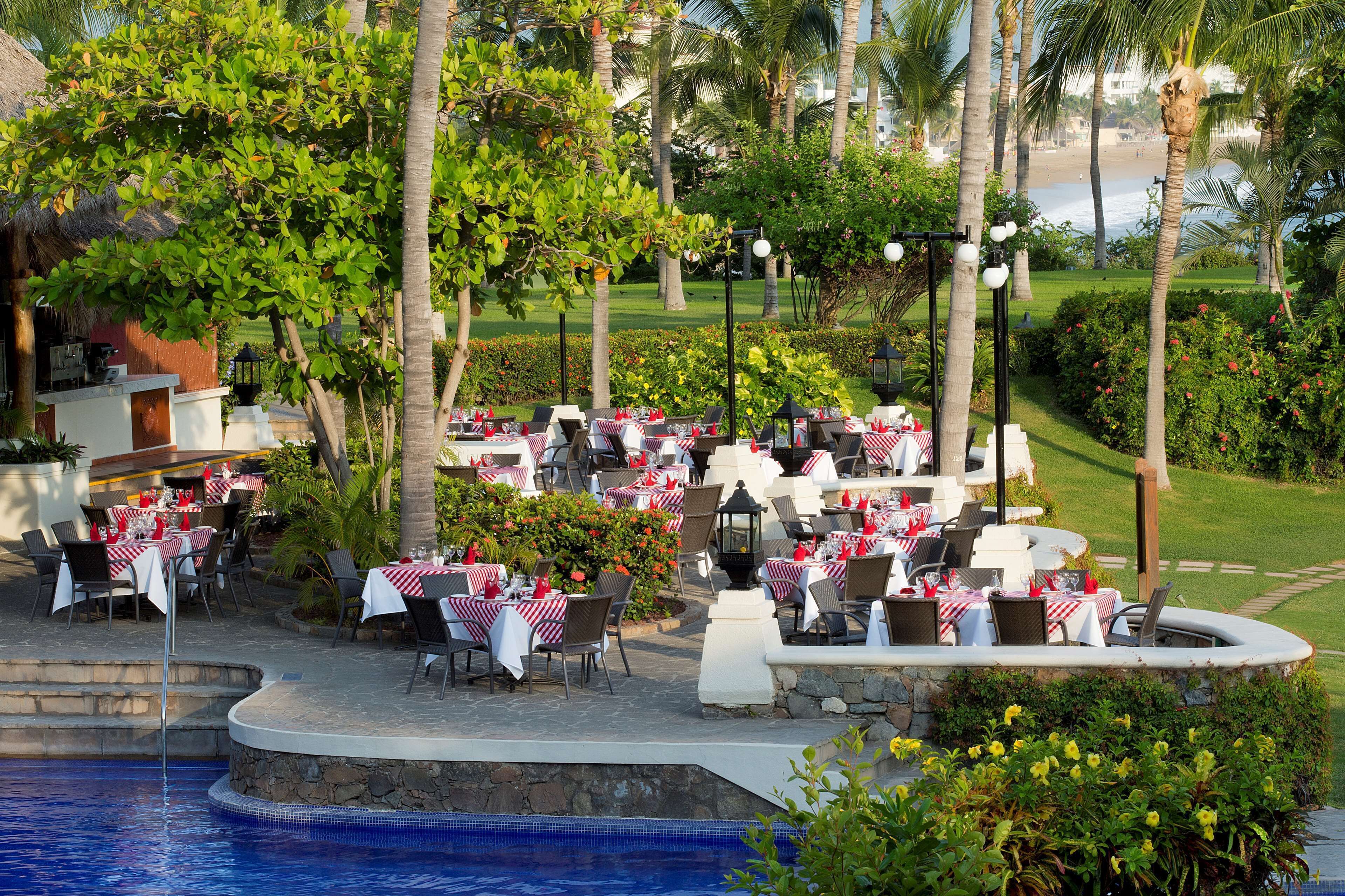 An outdoor dining area with tables set for guests, surrounded by lush greenery and a pool.