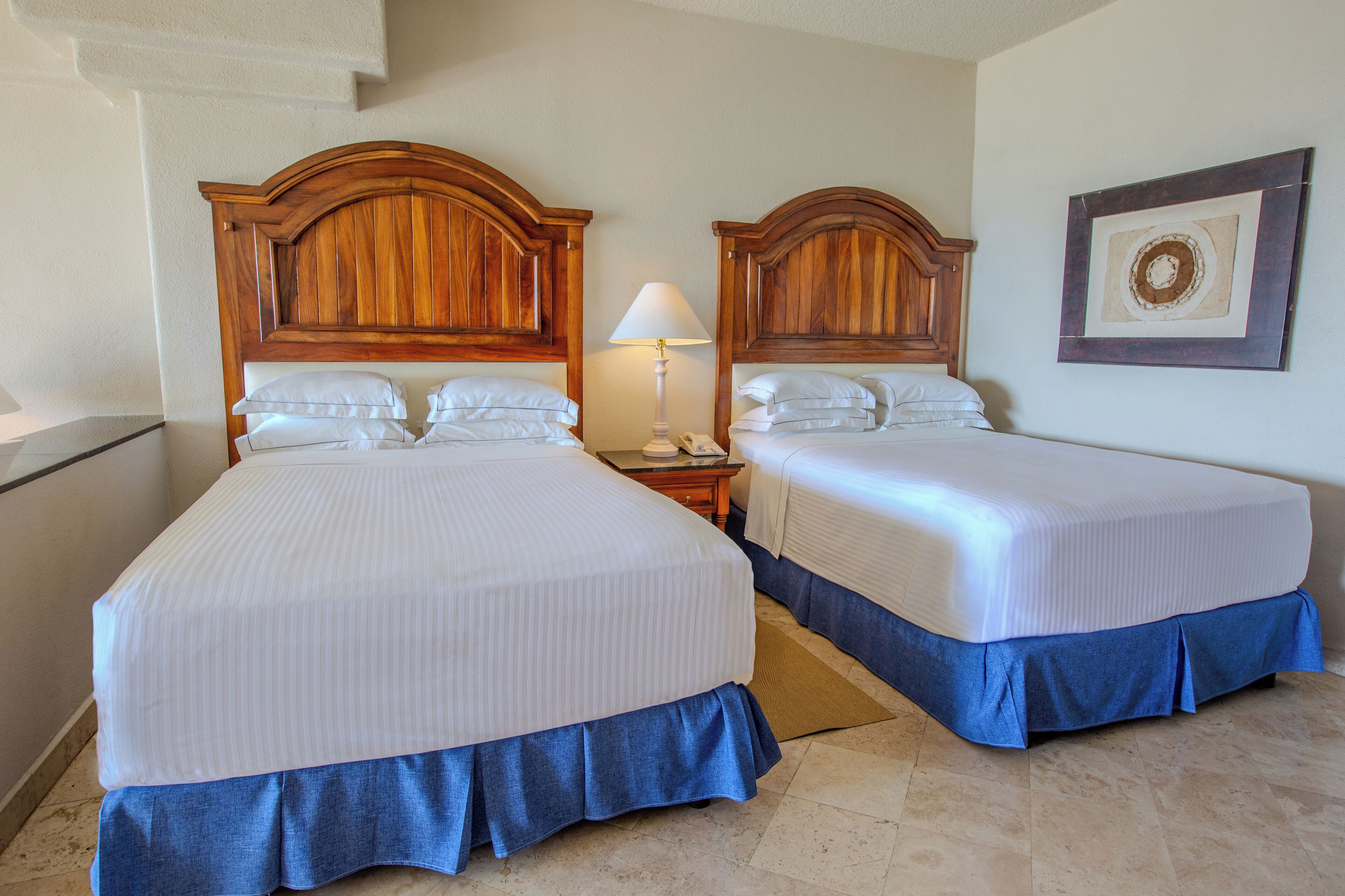 Una habitación de hotel con dos camas, una mesita de noche con lámpara y un cuadro enmarcado en la pared.