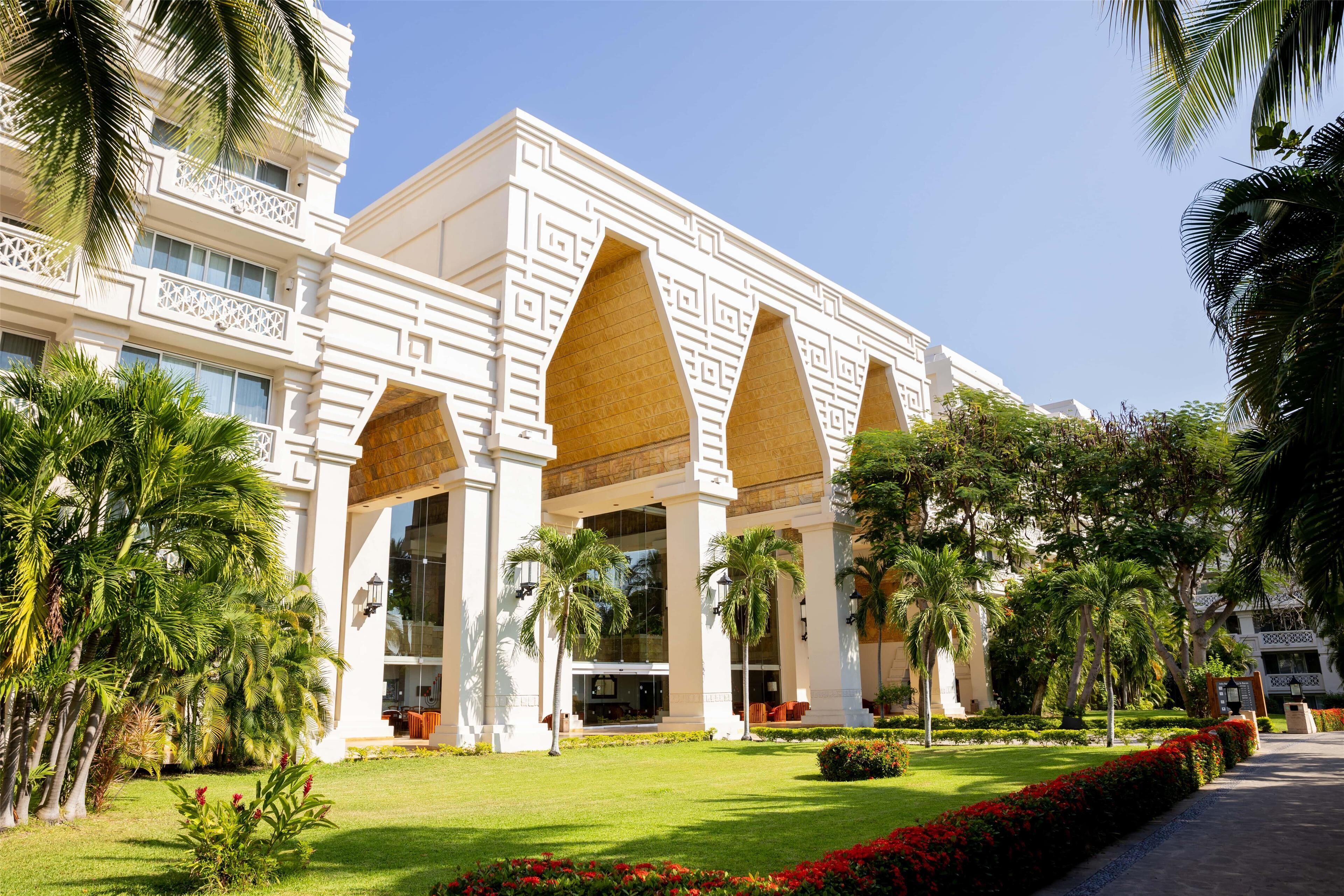 A hotel with a symmetrical facade, large arched windows, and a well-maintained garden with palm trees and red flowers.
