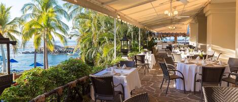 An outdoor dining area with tables set for guests, overlooking a beach with palm trees and a view of the ocean.