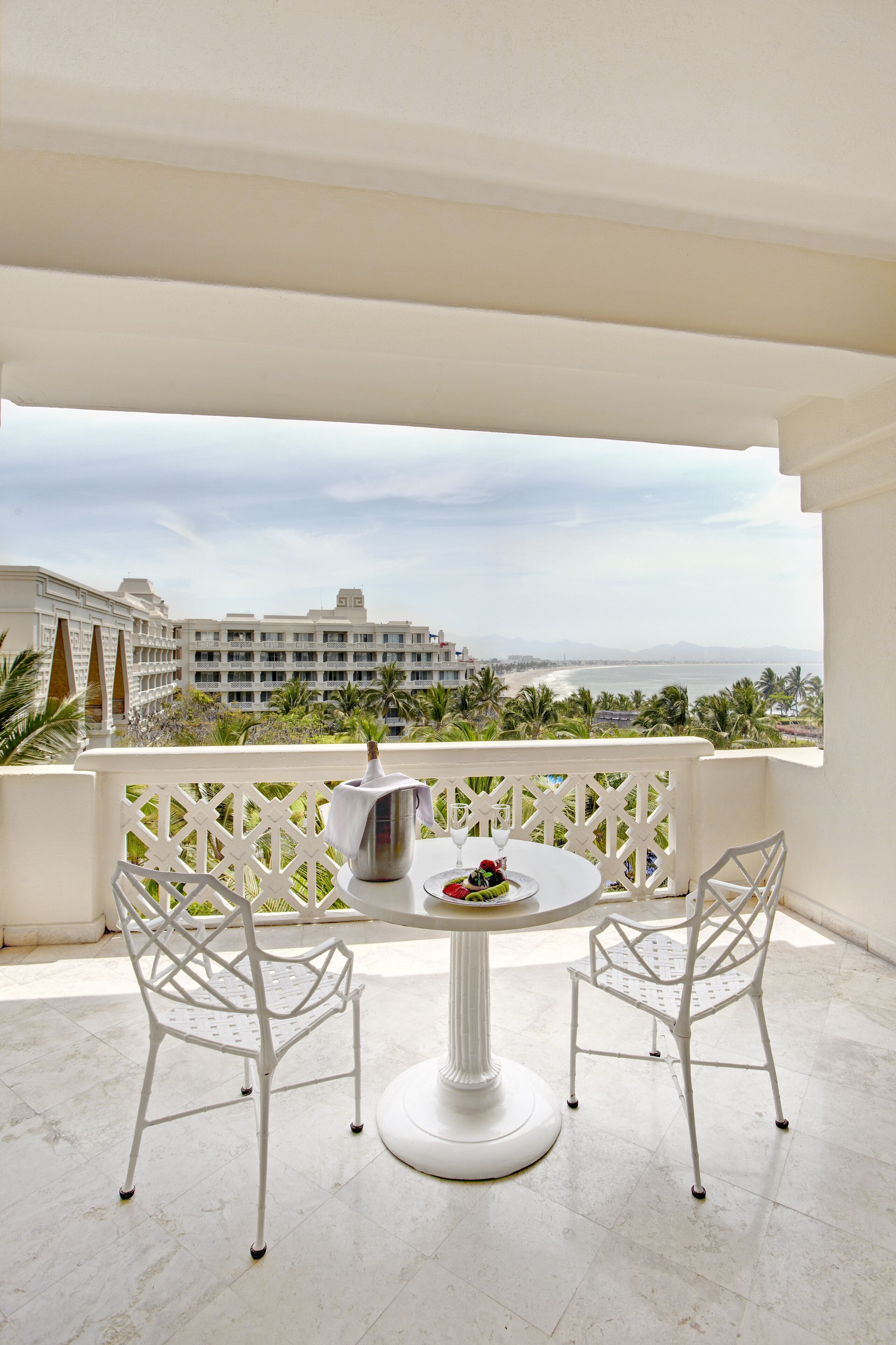 Un balcón con una mesa y sillas blancas, con vista a una playa y edificios de hotel.