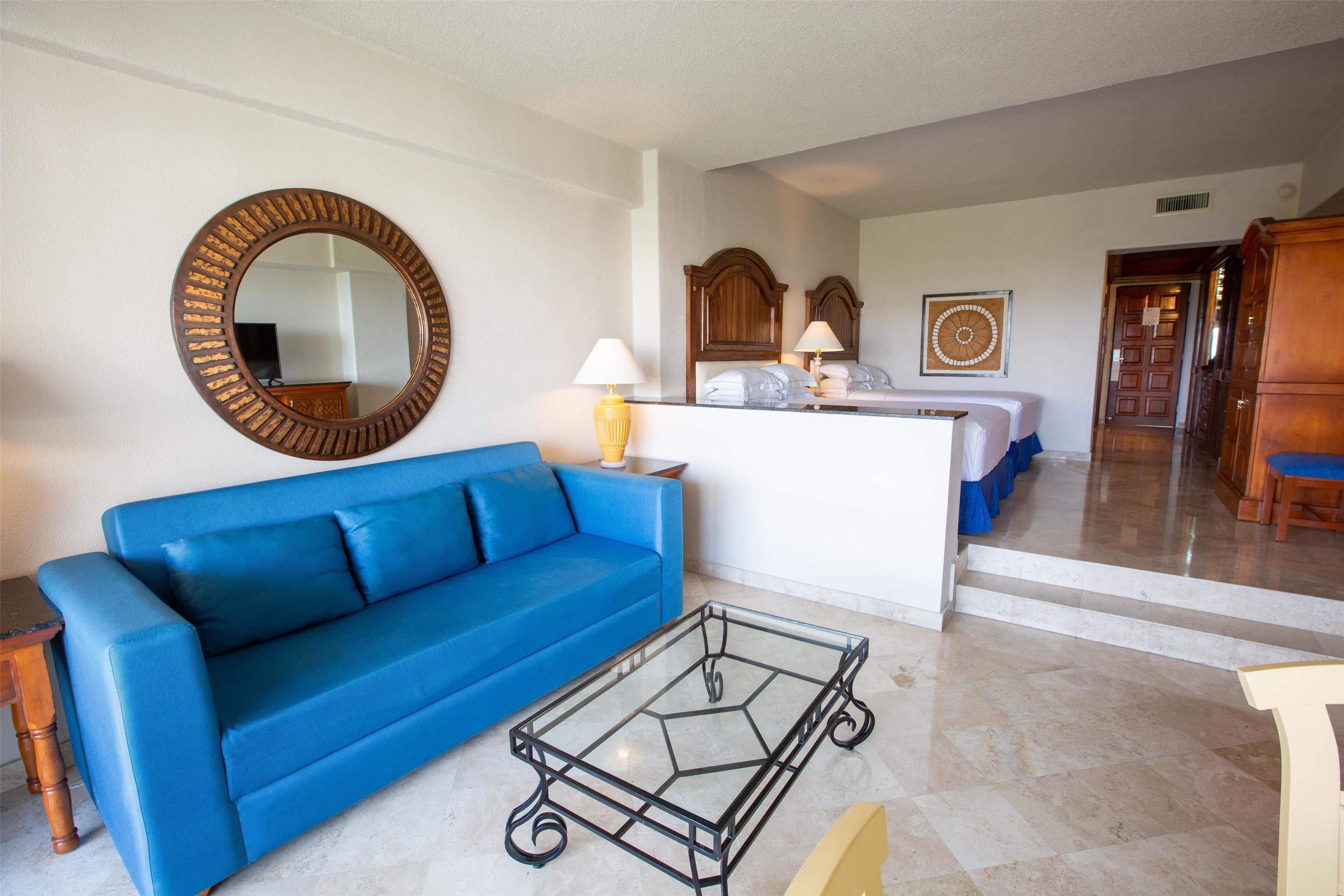 A hotel room with a blue sofa, a glass coffee table, a wooden headboard, a bed with a wooden headboard, a wooden wardrobe, and a mirror on the wall.