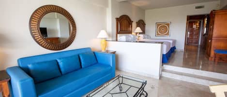 A hotel room with a blue sofa, a glass coffee table, a wooden headboard, a bed with a wooden headboard, a wooden wardrobe, and a mirror on the wall.