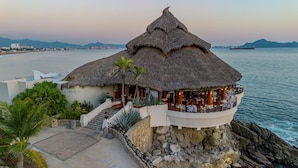 A thatched-roof structure with outdoor seating, overlooking a body of water and a distant cityscape. - Barceló Karmina All Inclusive (Manzanillo)