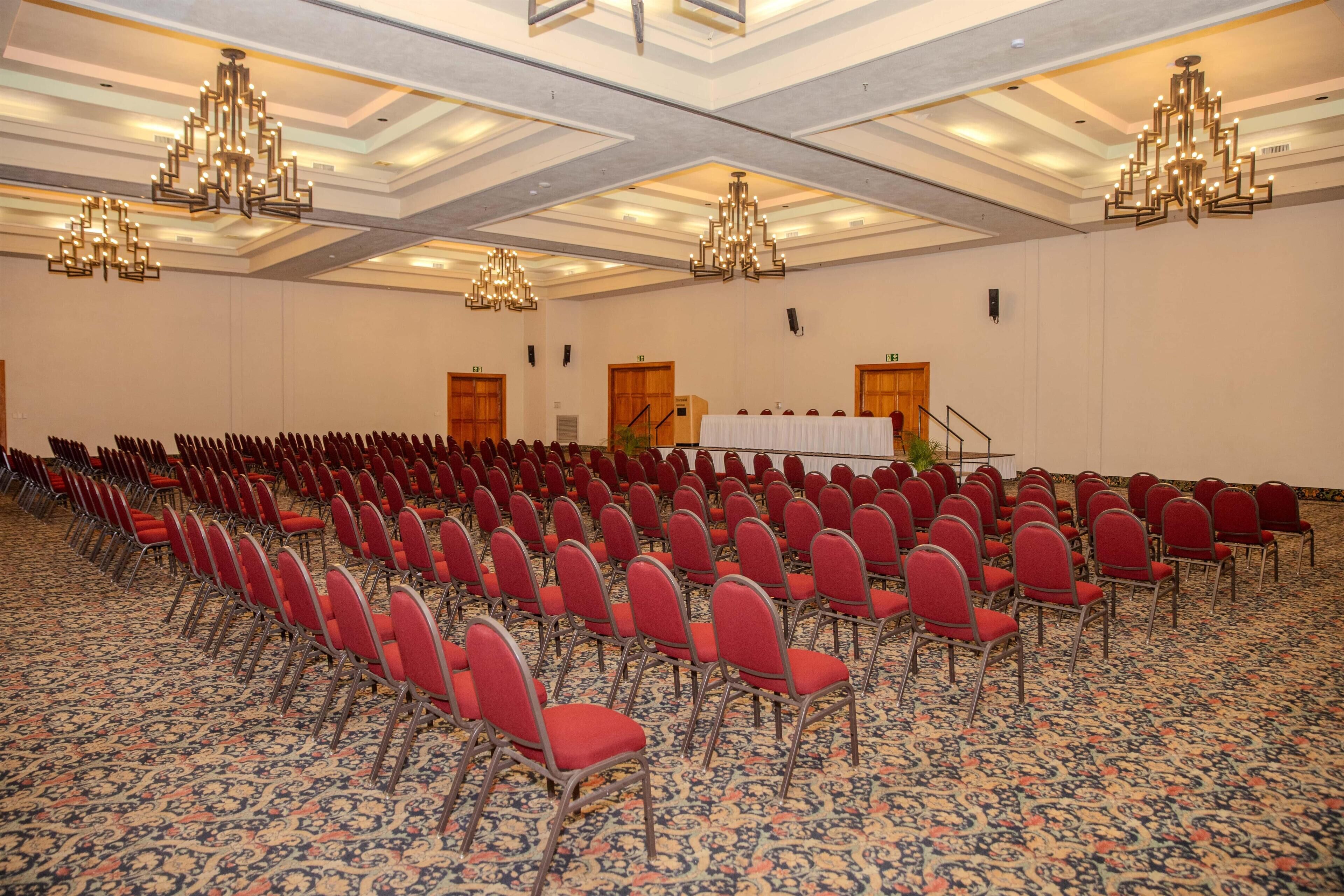 Un salón de conferencias con filas de sillas rojas, un escenario con una mesa blanca y candelabros.
