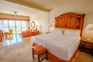 Habitación de hotel con una cama grande, cabecera de madera, zona de estar con una silla y una mesa, y un balcón con vistas.