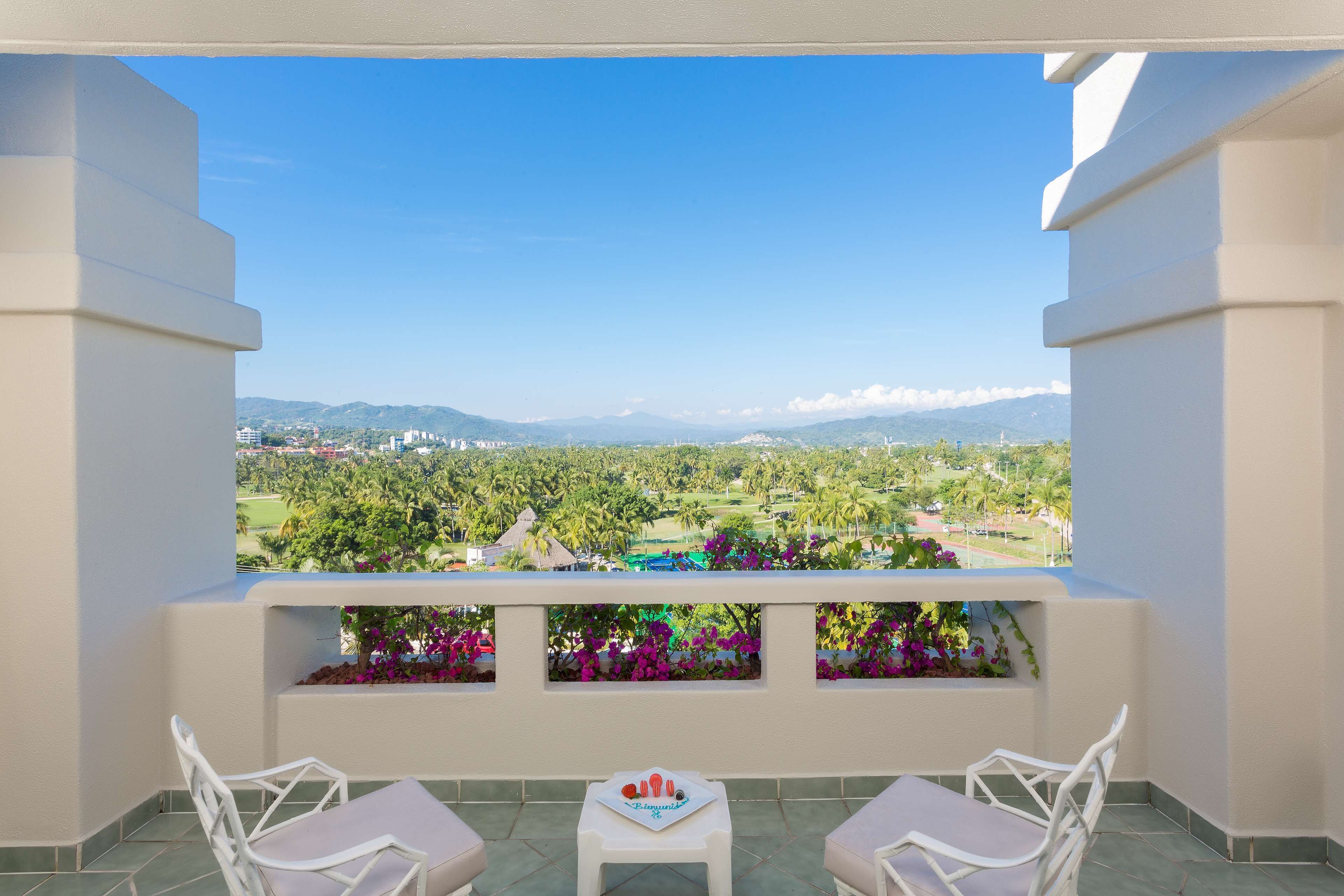 Un balcón con dos sillas blancas y una pequeña mesa, con vistas a un paisaje exuberante de montañas y vegetación.