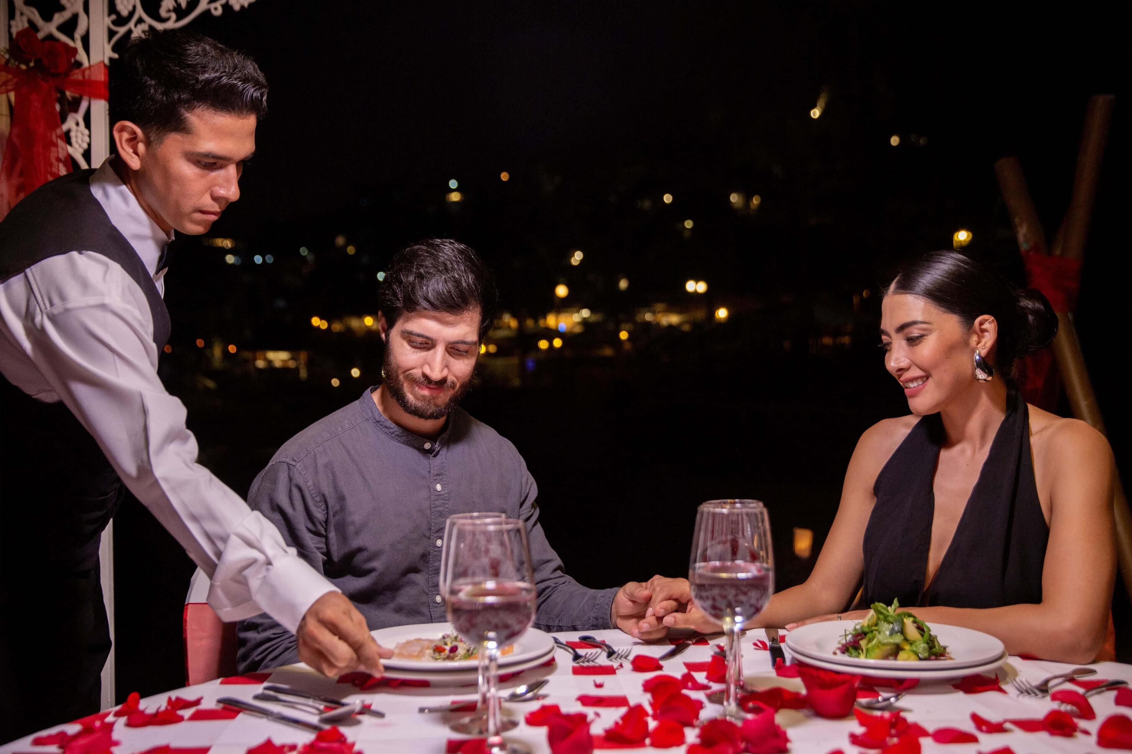 Un couple est assis à une table avec vue sur la ville, un serveur apporte les plats, et des pétales de rose rouge sont disposés sur la table.