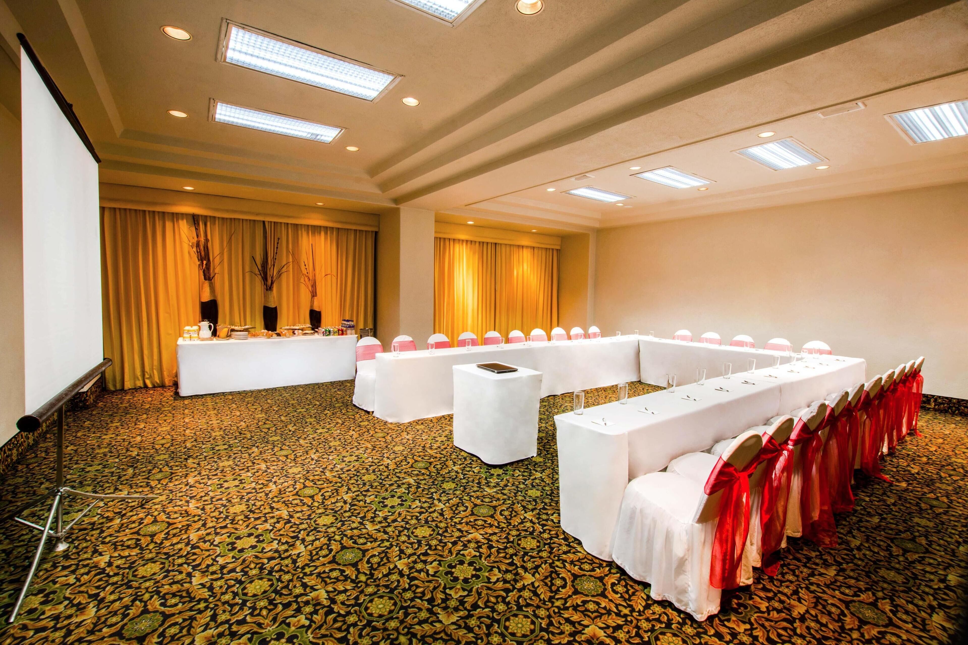 Un salón de conferencias con una larga mesa, manteles blancos y cintas rojas.