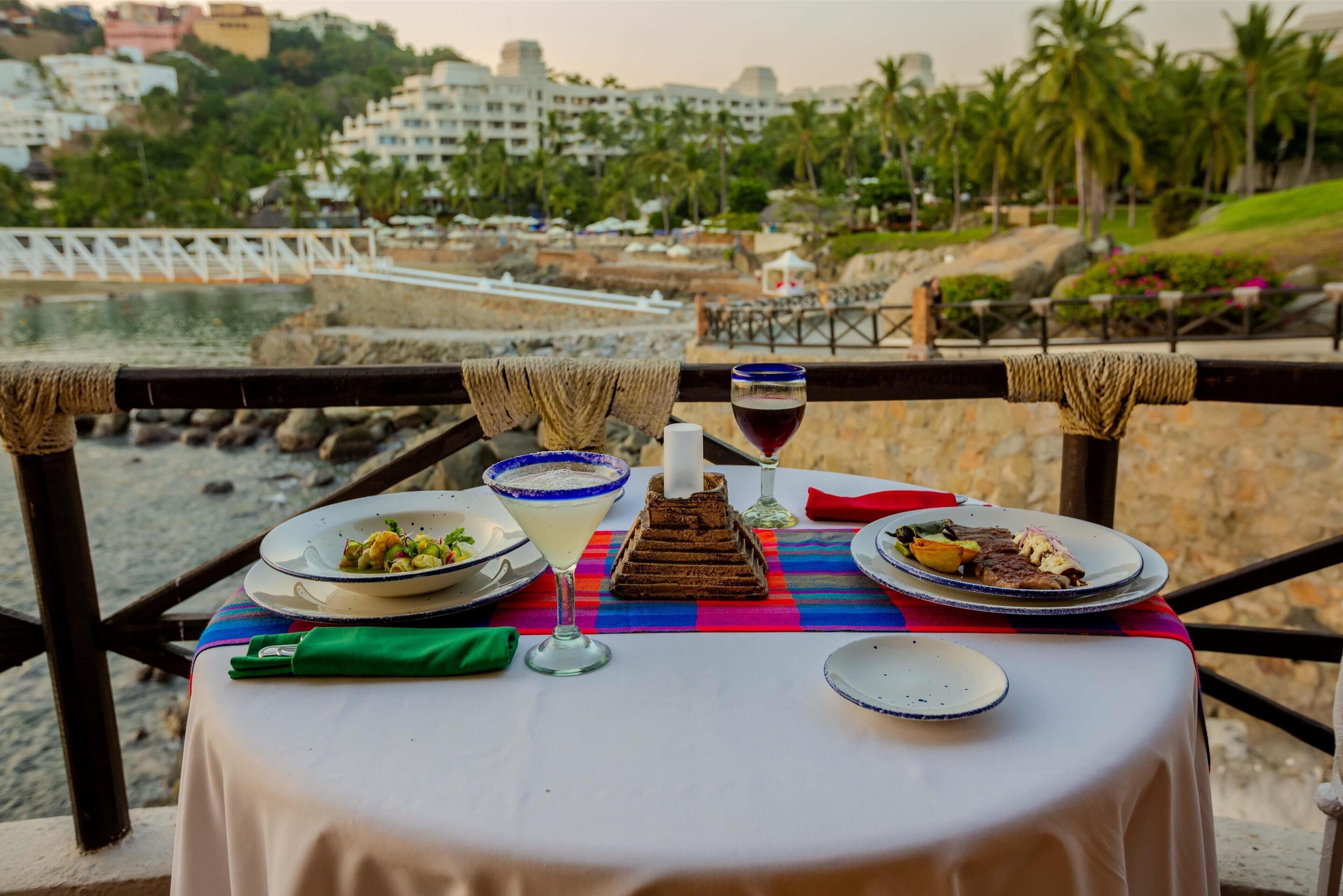 Une table dressée avec des assiettes remplies de nourriture, un verre de vin rouge et un centre de table décoratif, donnant sur une plage et des palmiers.