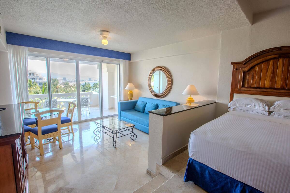 Quarto de hotel com cama, sofá azul, mesa de jantar com cadeiras, janela grande e varanda.