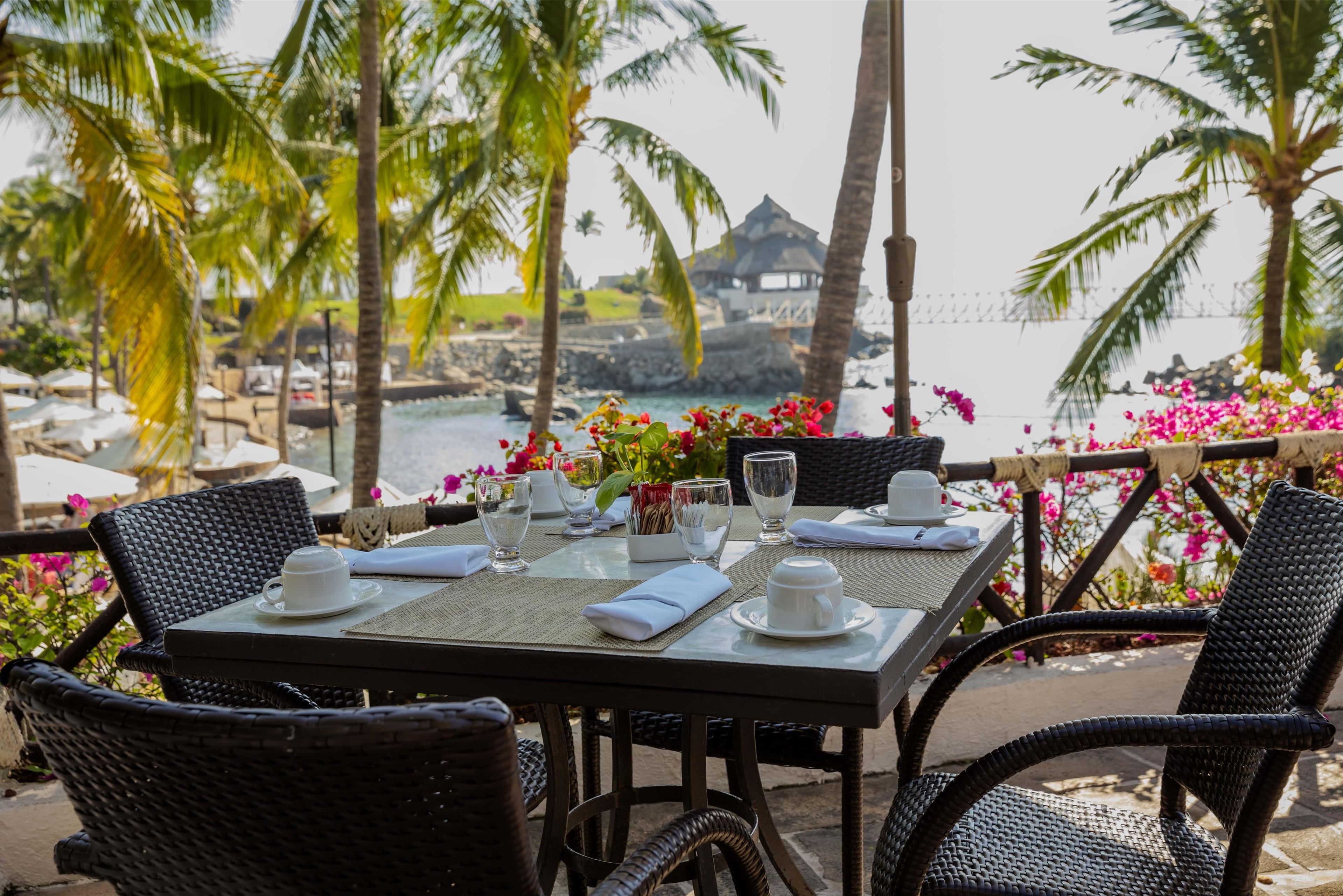 An outdoor dining area with wicker chairs and a table set for two, overlooking a beach with palm trees and a thatched-roof structure in the background.