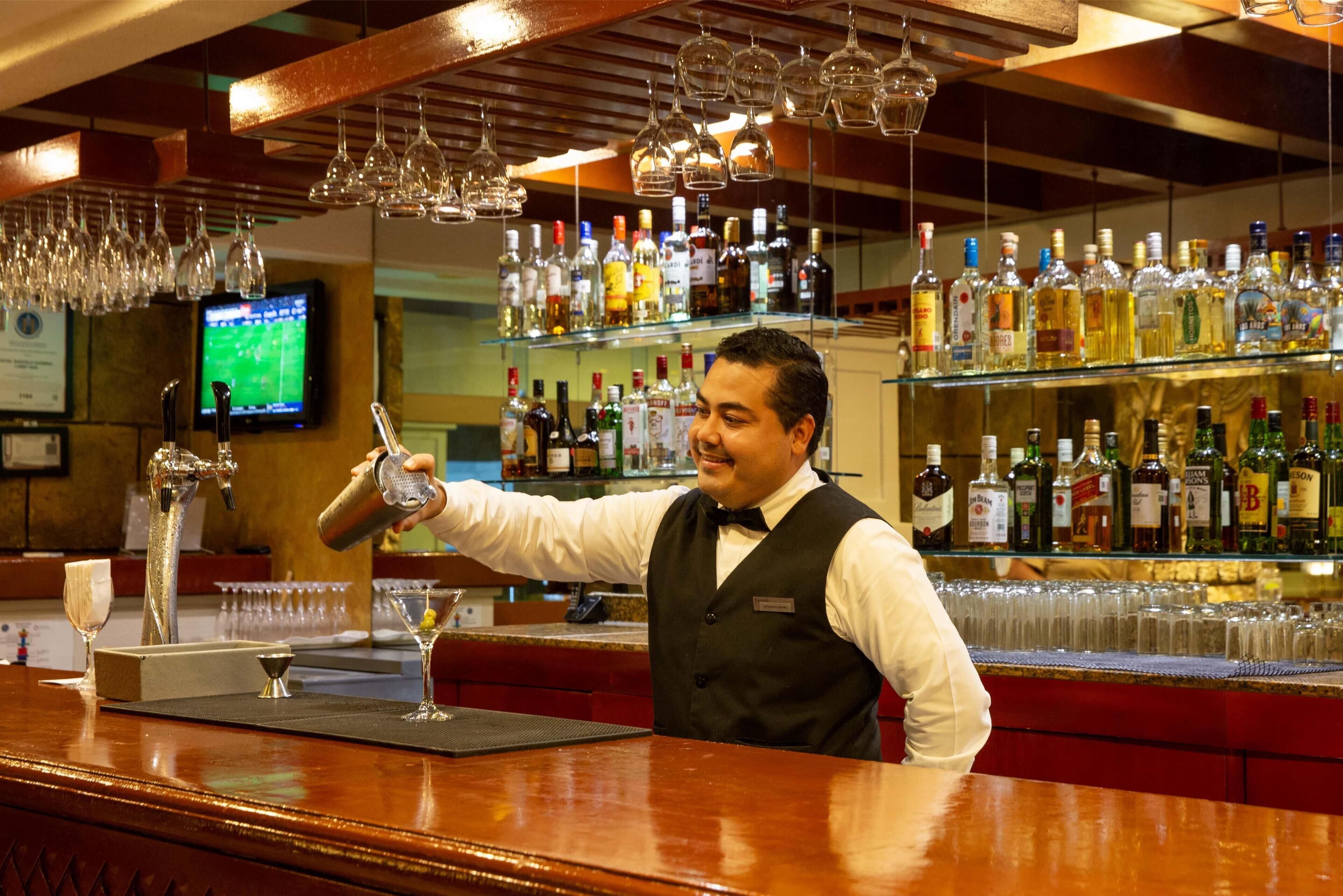 A bartender at a bar with a well-stocked liquor shelf and a television in the background.