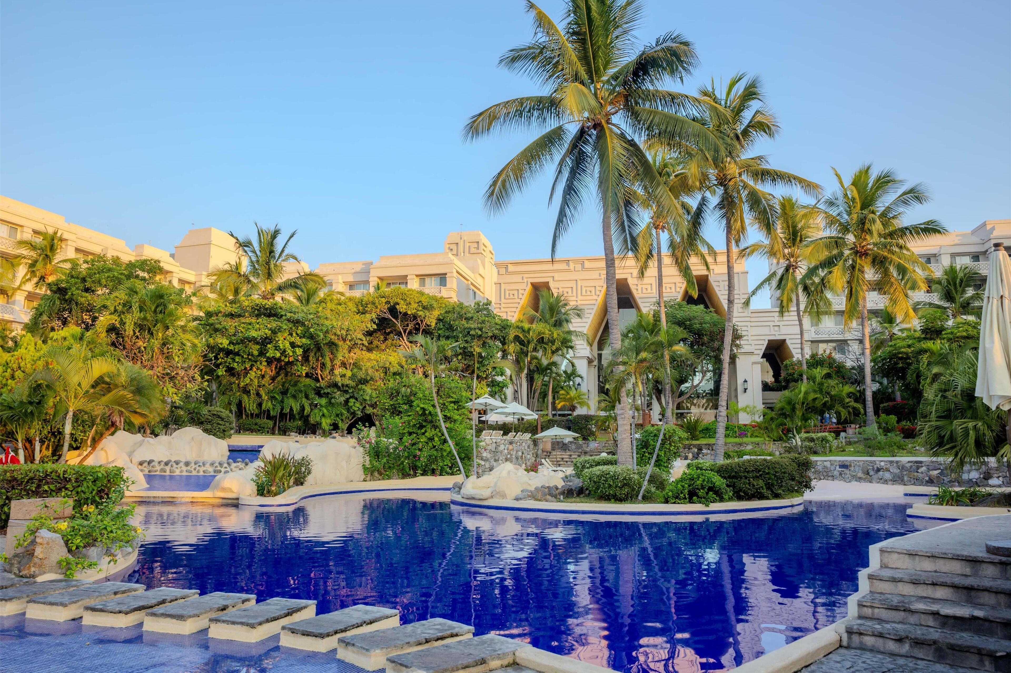 A pool with a stone border, palm trees, and a building with a geometric facade in the background.
