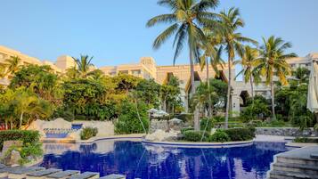 A pool with a stone border, palm trees, and a building with a geometric facade in the background.