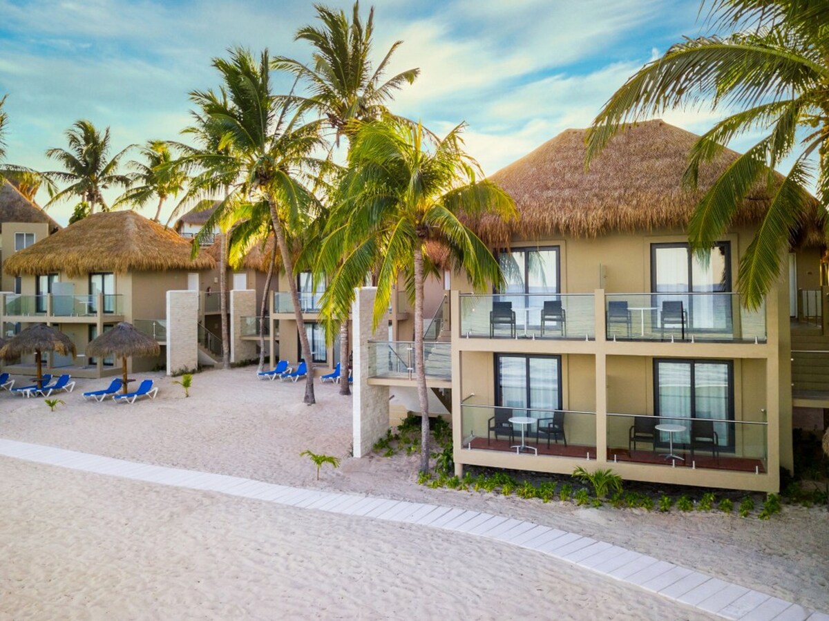 A four room villa surrounded by palm trees sits behind a sandy beach walking path.