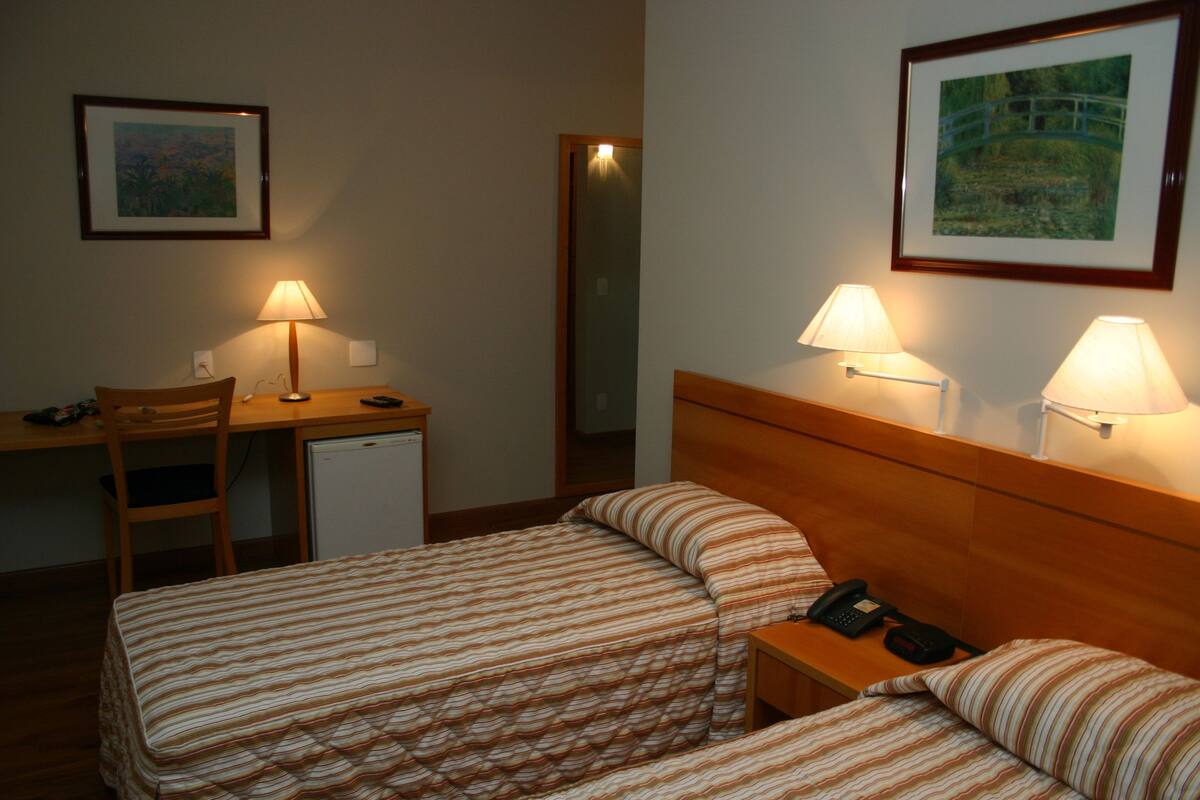 A hotel room with two beds, a desk, a chair, a small fridge, and two bedside lamps.