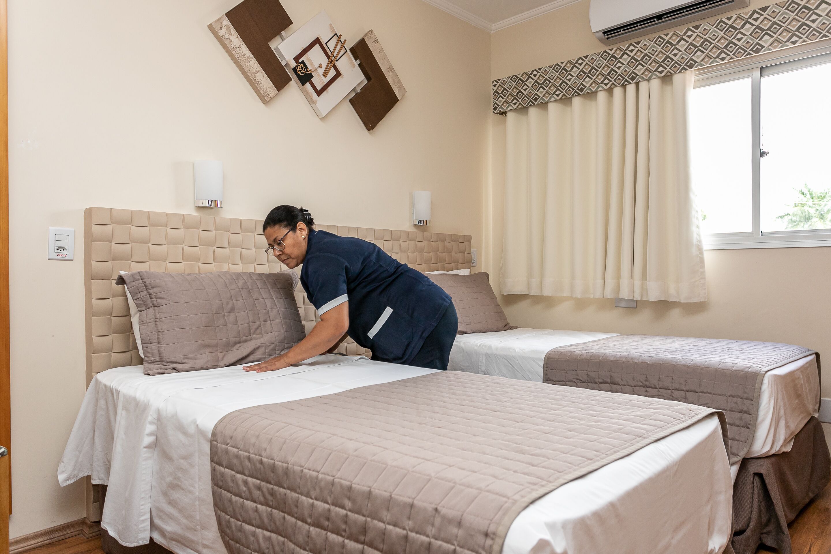 A hotel room with two beds, a beige headboard, a window with curtains, and a wall-mounted air conditioner.