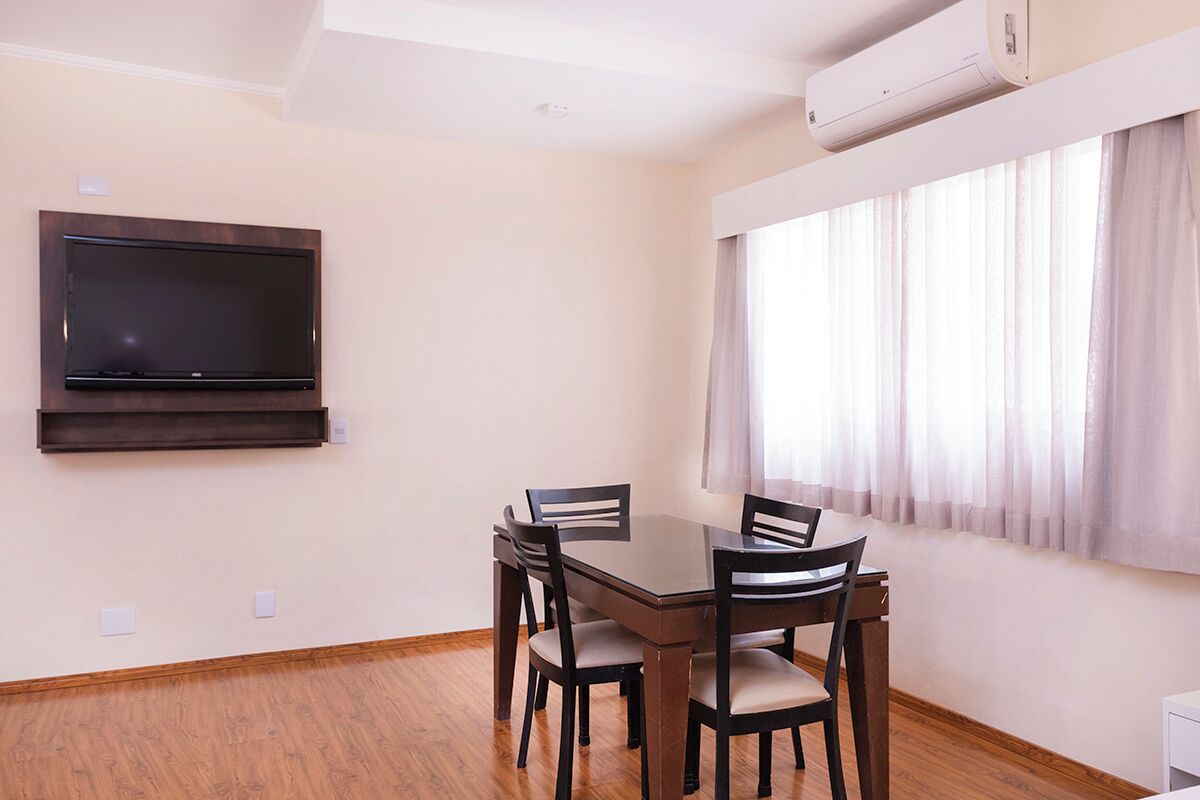 A dining area with a wooden table and chairs, a flat-screen TV mounted on the wall, and a window with sheer curtains.