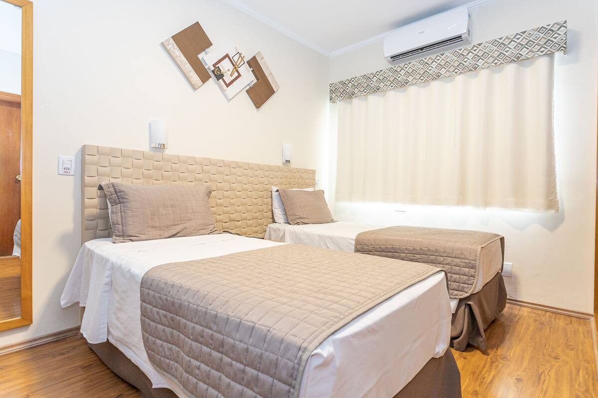 A hotel room with two beds, a wooden floor, a wall-mounted air conditioner, and a window with patterned curtains.