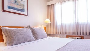 A neatly made bed with a quilted blanket and two pillows, a bedside lamp, and a window with sheer curtains. - Hotel Marbor (Mogi das Cruzes)