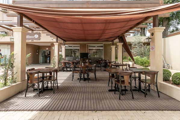 A covered outdoor seating area with tables and chairs, a sign for 'Hotel Marbor', and a brick building in the background. - Hotel Marbor (Mogi das Cruzes)