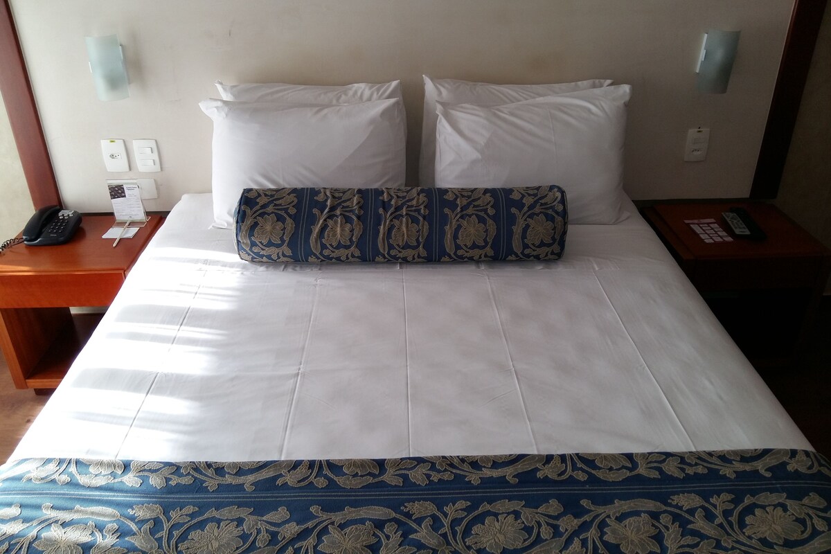 Habitación de hotel con una cama bien tendida, una mesita de noche con un teléfono y un control remoto, y una lámpara.