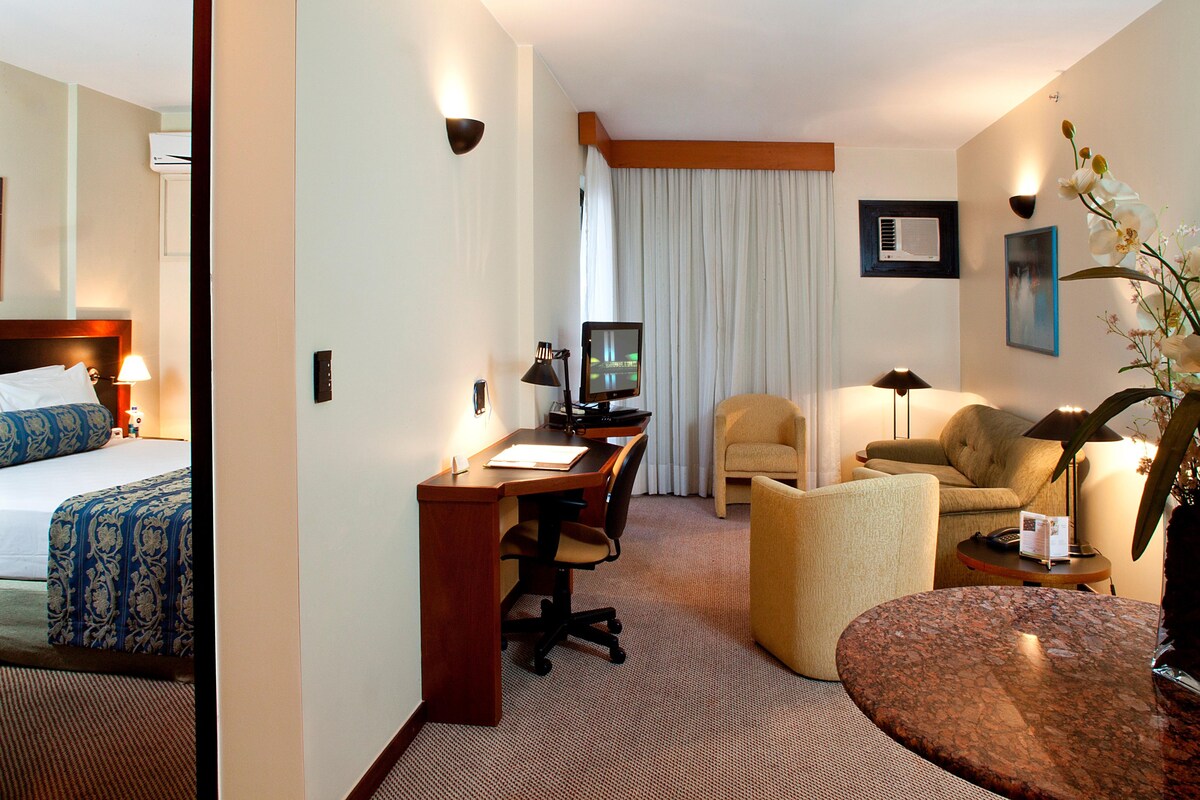 Habitación de hotel con cama, escritorio, silla, sofá y una pequeña mesa con un florero.