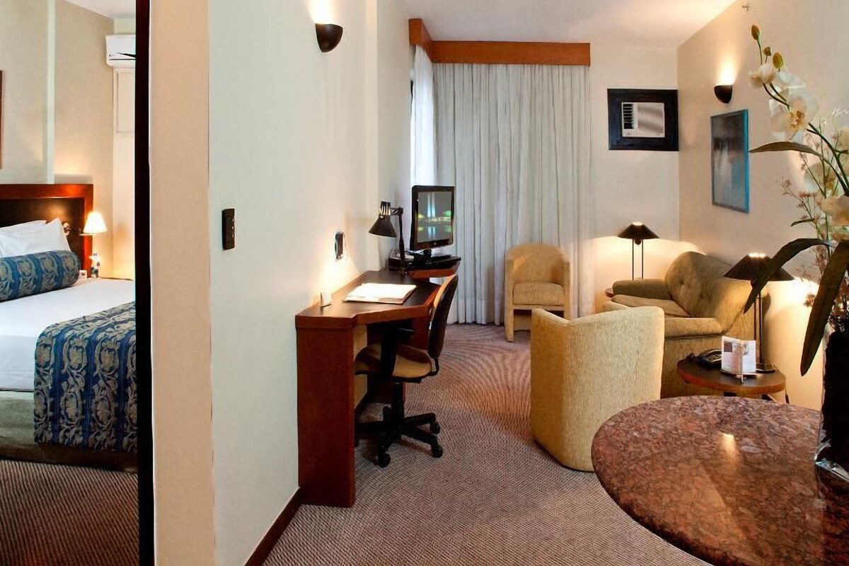 Habitación de hotel con cama, escritorio, dos sillas, una mesita y televisor.