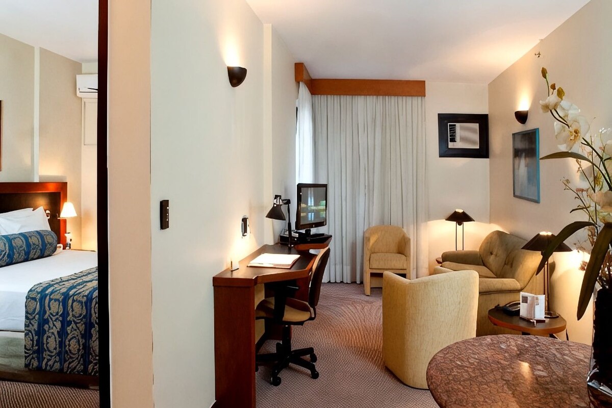 Habitación de hotel con cama, escritorio, computadora, dos sillones, una mesita y una planta en maceta.