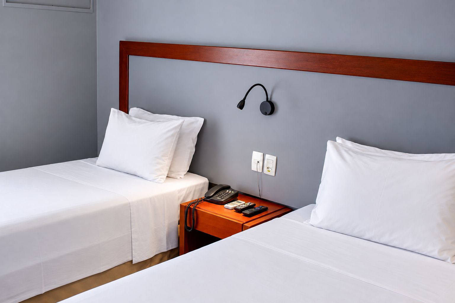 A hotel room with two single beds, a wooden headboard, a bedside table with a telephone and remote controls, and a wall-mounted light.